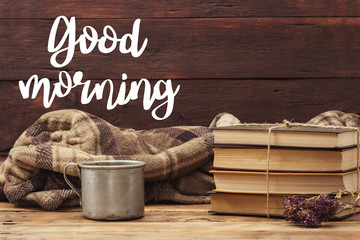 A metal cup with hot tea, coffee, a warm blanket, a pile of books tied with a rope, a pot of hips on a wooden table. Added text Good Morning