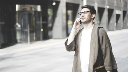 A hipster guy wearing glasses and a khaki jacket talking on his smartphone and laughing in the street. Portrait shot