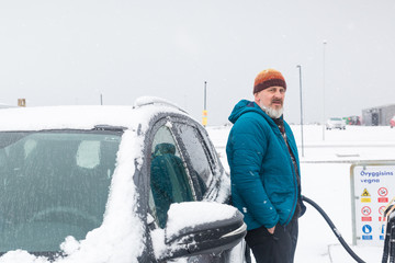 Mann mit blauer Jacke lehnt beim Tanken im Schnee am Auto