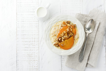 Oatmeal porridge with caramelized persimmon and walnuts in a light ceramic bowl on a white wooden background. Selective focus. Top view. Copy space.