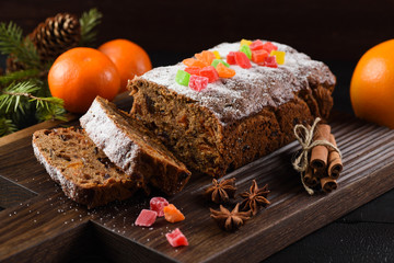 Traditional sliced freshly baked fruit cake decorated with candied fruits, star anise, cinnamon and clementines on dark background