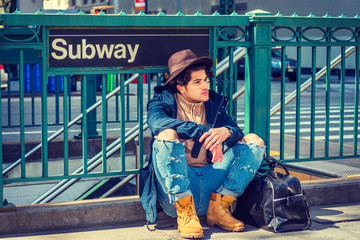 Young American Man with freckle face, curly hair, traveling in New York,  wearing blue jacket, jeans, yellow boot shoes, Fedora hat, black leather bag on ground, sitting on street by Subway sign..