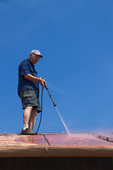Man is washing the roof with a high pressure washer. Silhouette of a worker cleaning a red metal sheet roof by water.