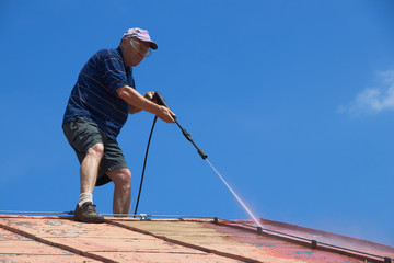 Man is washing the roof with a high pressure washer. Silhouette of a worker cleaning a red metal sheet roof by water.