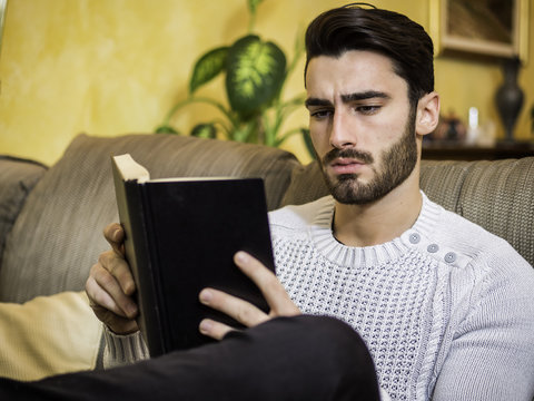 Handsome black hair young man reading book at home, sitting on couch