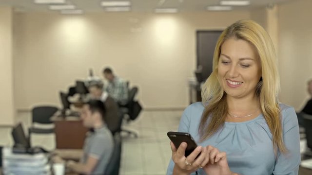 Closeup portrait of enjoyed blonde women speaking on phone. She smiling to side. Blue solid background. Modern office.