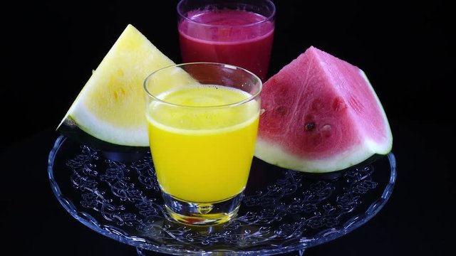 Pyramids of red and yellow water melon and couple of glass with freshly squeezed juice of them, rotating on black background against the clock close up. Vibrant natural texture in UHD 4k, 3840x2160.