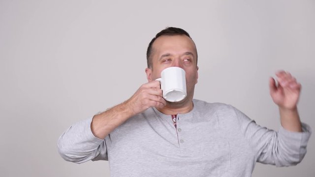 Man sleepy tired wearing nightwear holding coffee mug about to drink. Male having hard morning, getting energy, on grey. Slow motion