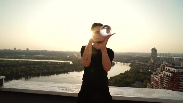 Young man shows contact juggling on roof of skyscraper high above the city at sunset 4K