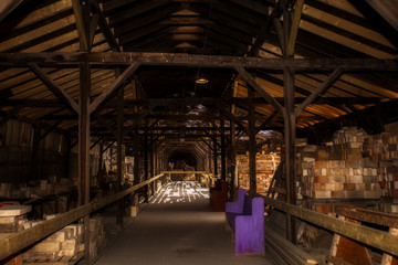 Wood shell of rafters and beams dotted with sunlight storing stacks of bricks between a hallway of a discontinued brick factory