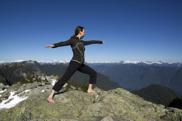 Middle Aged Caucasian Woman Holding Hatha Yoga Warrior Pose on Winter Snowcap Sunshine Mountain Peaks with Blue Sky Background