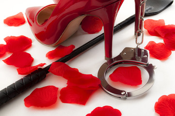 Kinky sex and BDSM games concept with a pair of red and shiny high heel stilettos, a leather crop under the shoes, a pair of metal handcuffs and rose petals surrounding the scene