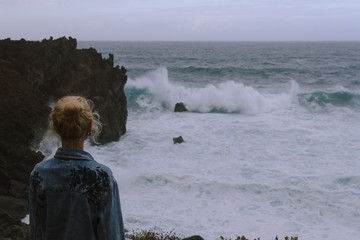 Young woman standing next to the ocean and looking at rough sea and breaking waves