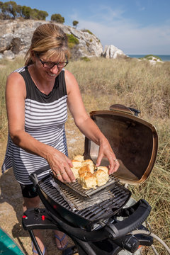 Mature Woman Cooking An Omelette On An Outdoor Portable Barbeque Near A Beach.