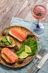 Snack with red fish and avocado served on a wooden plate, pink champagne in a glass, spoon fork, blue napkin, wooden brown background.