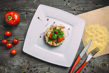 Couscous with green peas, peppers, zucchini and ginger on square white plate with cutlery. Wooden gray background and clean paper, top view