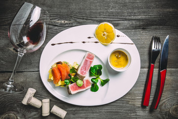 Tuna steak with tartar sauce, grapefruit, peach, dried tomatoes and a glass of red wine. On the white round plate with cutlery, on a wooden old table, top view