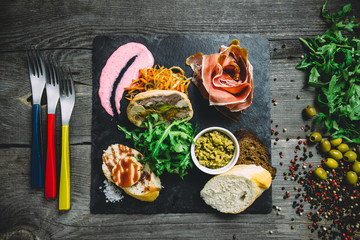 Jamon, meatloaf with mushrooms, bread, arugula, sauce, olives and carrots on a black square plate with forks. Wooden gray background, top view