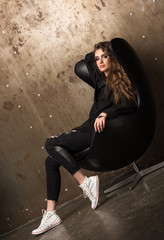 Young beautiful woman sitting in the black chair. The casual look, black shirt, jeans and white sneakers