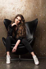 Portrait of beautiful young woman sitting in the black chair. Contemporary appearance. Black clothes and white sneakers