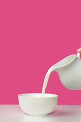 cropped shot of hand pouring fresh milk from jug to bowl on pink