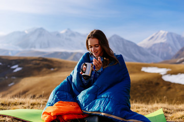 smiling young girl sitting in a blue sleeping bag drinking hot tea on the background of the Caucasian mountains - 186959417