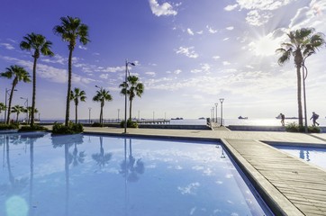 Molos Promenade and skyline of the coast in Limassol city in Cyprus at sunrise. View of the boardwalk pier path landmark with palm trees, pools of water, the Mediterranean sea and people walking.