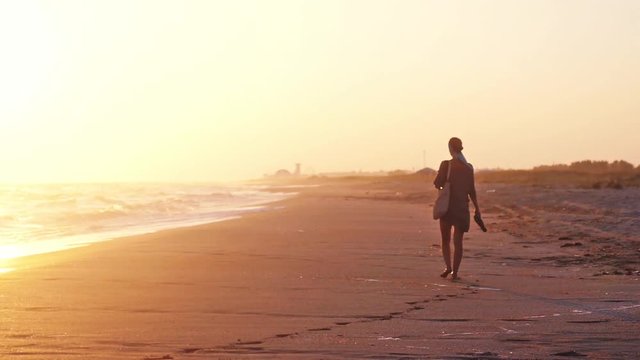 Woman with a bag and beach shoes in hands walking on beach at amazing sunset in summer in slow motion. 1920x1080