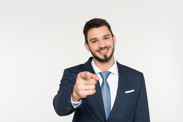 smiling young businessman pointing at camera isolated on grey