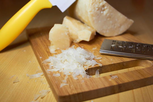 3 different pieces of coarse Italian Parmesan cheese, a special cheese knife for Parmesan, enters the cheese head at an angle, a small iron grater for cheese, grated parmesan flakes on a wooden board.