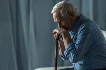 Senior sad man leans on a cane while sitting on sofa - obrazy, fototapety, plakaty
