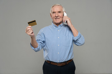 Confident senior man talking on phone and holding credit card isolated on gray background