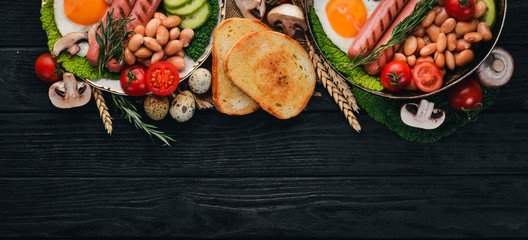 Full English breakfast with fried eggs, sausages, beans, toasts and coffee on copy space background. On a black wooden background.