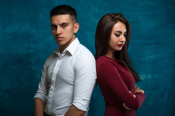 Young couple - serious charming woman and manly man stand with their backs to each other with hands clasped on chest on blue background