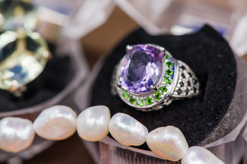 Macro shot of silver engagement ring in gift box next to pearl necklace on colorful, sparkling background. Ring made of sapphire, amethyst, chrome diopside stones. Healing, powerful, natural crystals 