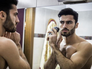 Handsome young man using shaving machine at the mirror. 