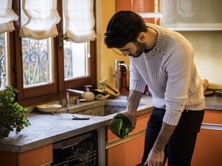 Young handsome man putting green bowl into dishwasher in kitchen at home.