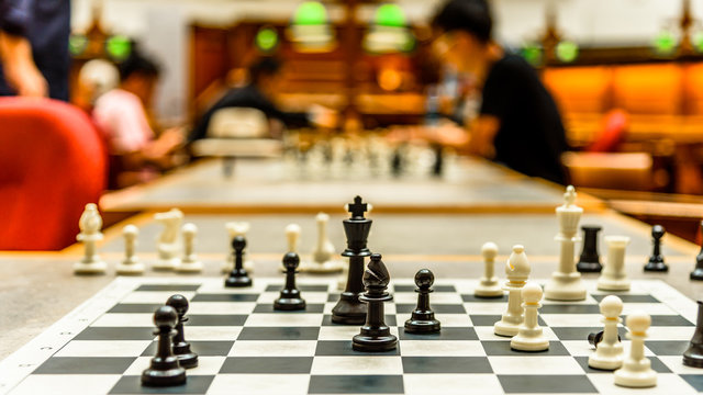 A completed game of chess with black and white pieces, a fallen king piece and people still playing in the background.
