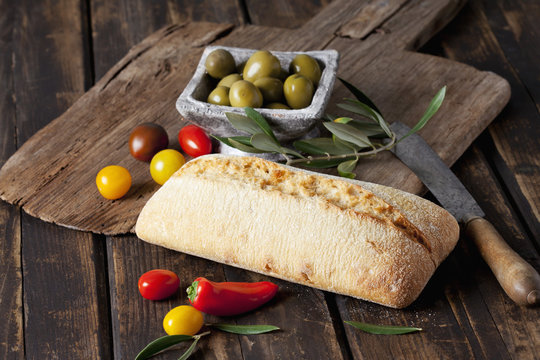 Ciabatta bread with green olives in bowl, tomatoes and mini capsicum on wood