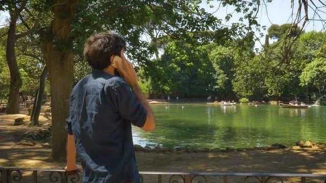 Young Man talking on smartphone on phonecall by a Pond in villa borghese park in Rome on sunny summer day slow motion steadycam