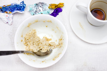 An overhead photo of dirty plate with porridge leftovers, chocolate candies wrapper, spoon and a cup of black espresso coffe.