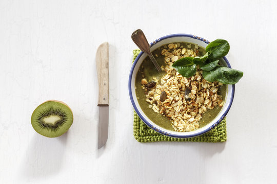 Bowl of green smoothie with mix of oat flakes and granola, kitchen knife and half of kiwi