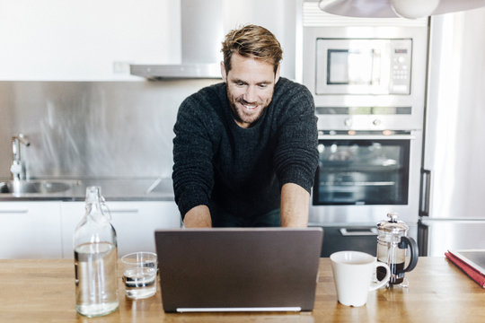 Portrait of smiling man standing in kitchen using laptop