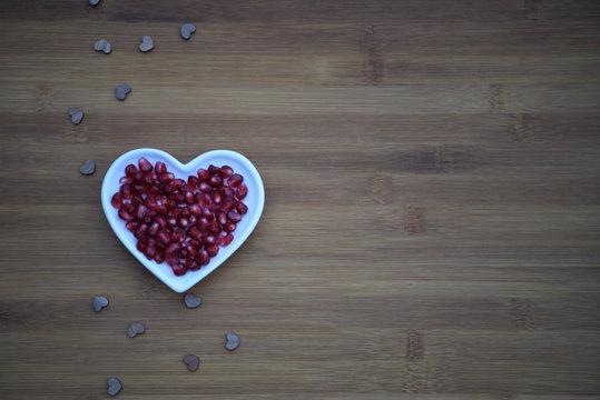 healthy food photography image of ripe juicy shiny red pomegranate seeds in a white love heart shape dish on a natural dark wood background with copy space