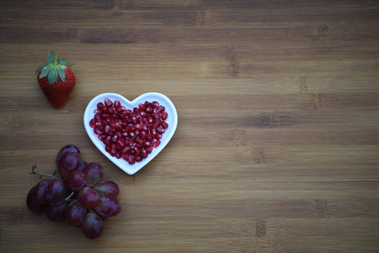food photography image with red shiny pomegranate seeds in a white love heart shape dish with a strawberry and bunch of fresh purple grapes on a dark natural wood background with copy space