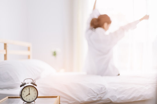 Close up alarm clock. Happy woman stretching on bed after waking up in the early morning at window side. Have a good day.