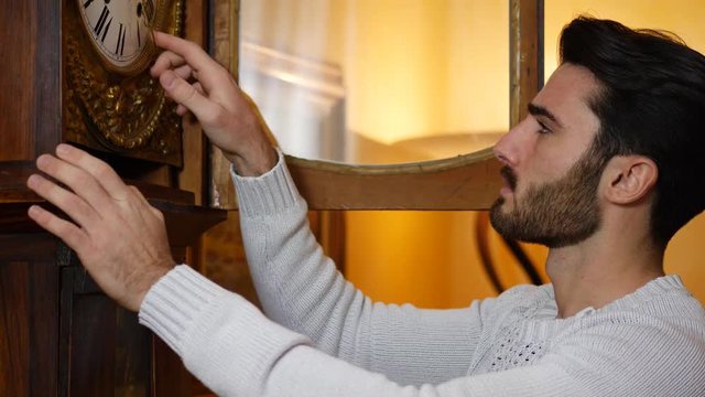Side view of young handsome man standing and setting vintage pendulum clock.