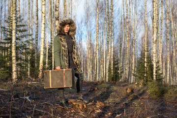 Young man in a green winter parka in a forest with a suitcase. © Kertu