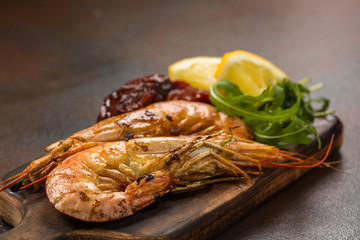 Fried tiger shrimps or langostinos with lemon, arugula, sun dried tomatos on a dark wooden cutting board. Placed on a background of dark table