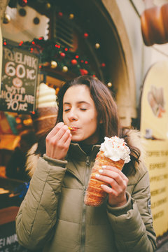 A beautiful girl in a warm jacket eats trdelnik or Trdlo with cream in her hands, in the winter in the Czech Republic, Prague at the Christmas market